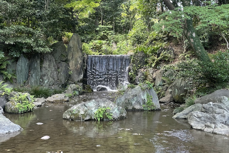 Kleiner Wasserfall im Park mit Steinbrocken im Vordergrund und grüner Vegetation im Hintergrund